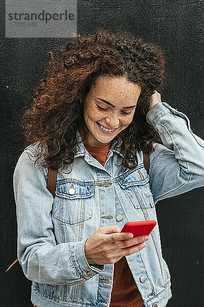 Lächelnde junge Frau  die eine Textnachricht über ihr Smartphone sendet  während sie mit der Hand im Haar steht