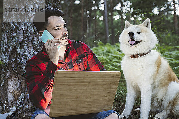 Mann mit Laptop  der mit seinem Handy telefoniert  während er neben einem Hund im Wald sitzt