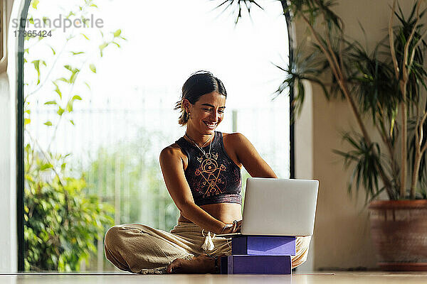 Lächelnde Frau  die einen Laptop benutzt  während sie im Schneidersitz im Fitnessstudio sitzt