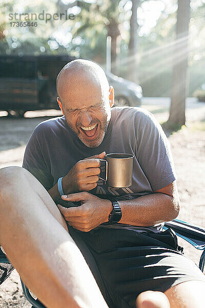Unbekümmerter Mann mit Kaffeetasse lachend an einem sonnigen Tag im Urlaub