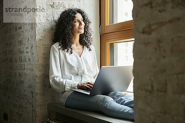 Geschäftsfrau  die mit ihrem Laptop auf der Fensterbank sitzt und träumt
