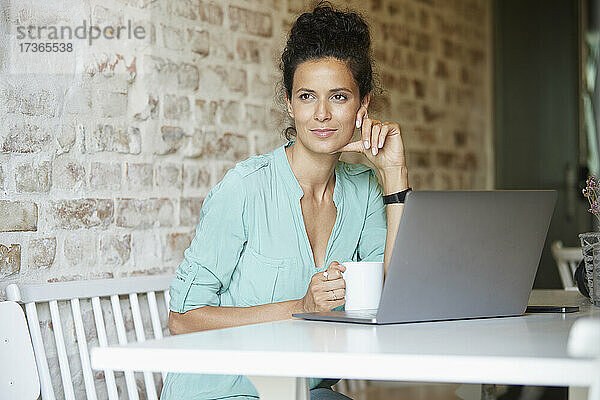 Nachdenkliche Frau sitzt mit Kaffeetasse und Laptop am Schreibtisch am Arbeitsplatz