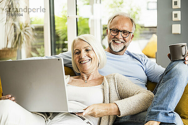 Frau lächelt bei Videoanruf über Laptop  während sie mit einem Mann auf dem Sofa sitzt