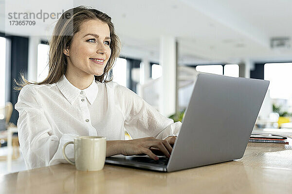 Lächelnde Geschäftsfrau mit Laptop auf dem Tisch im Büro