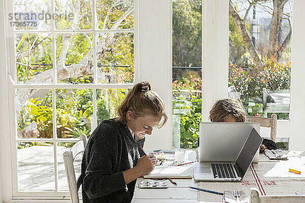 Teenager-Mädchen malt mit Wasserfarben an einem Tisch  und ein Junge an einem Laptop