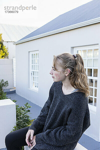 Teenager-Mädchen sitzt vor einem Haus auf einer niedrigen Mauer und wartet