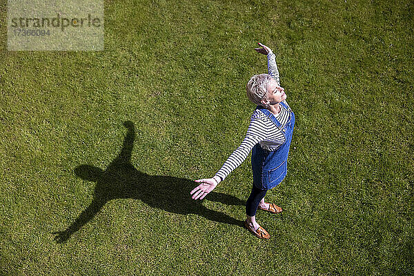 Ältere Frau mit ausgestreckten Armen im Gras an einem sonnigen Tag