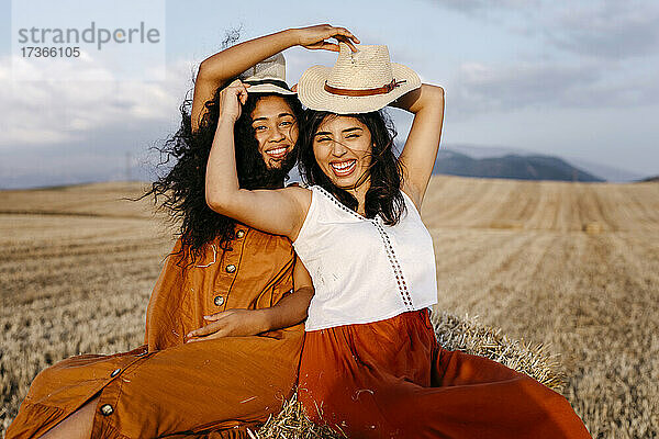 Freundinnen halten sich gegenseitig den Hut  während sie auf dem Bauernhof sitzen