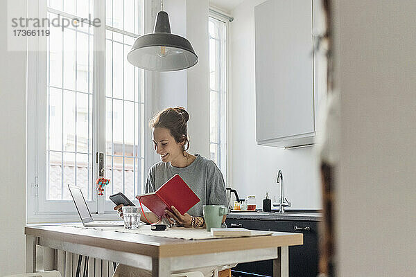 Lächelnde Freiberuflerin  die ein Mobiltelefon benutzt  während sie mit einem Buch in der Küche sitzt