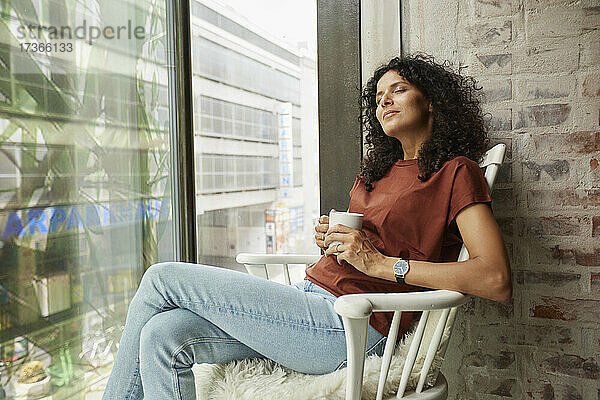 Entspannte Geschäftsfrau  die eine Kaffeetasse hält und mit geschlossenen Augen auf einem Stuhl am Fenster sitzt