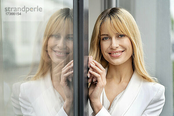 Lächelnde blonde Frau mit Spiegelung auf dem Fensterglas