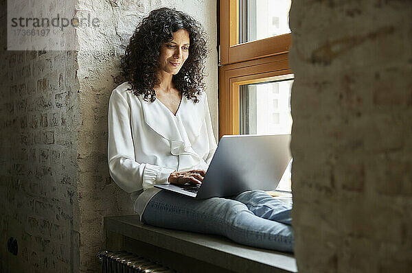 Lächelnde Geschäftsfrau  die einen Laptop benutzt  während sie auf der Fensterbank im Büro sitzt