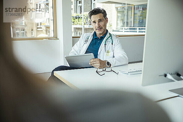 Männliche Fachkraft mit digitalem Tablet  die einen Patienten am Schreibtisch im Büro aufklärt