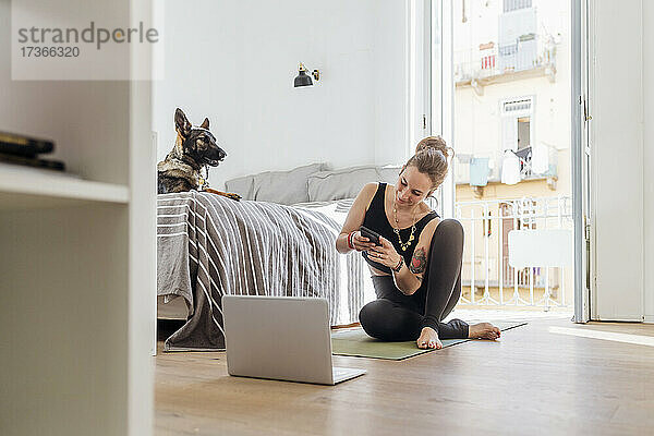 Frau  die ein Mobiltelefon benutzt  während sie auf einer Matte neben einem Laptop im Schlafzimmer einer Wohnung sitzt