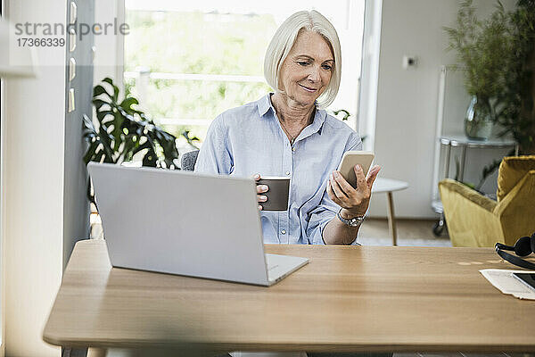 Reife weibliche Berufstätige  die ein Smartphone am Schreibtisch im Home Office benutzt