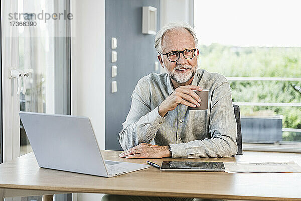 Geschäftsmann  der wegschaut  während er eine Kaffeetasse am Schreibtisch im Heimbüro hält