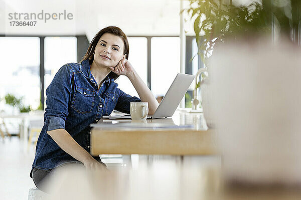Geschäftsfrau lehnt sich mit ihrem Laptop im Büro an einen Tisch