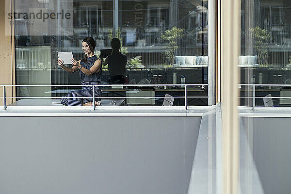 Lächelnde Geschäftsfrau  die ein digitales Tablet benutzt  während sie auf dem Balkon eines Büros sitzt