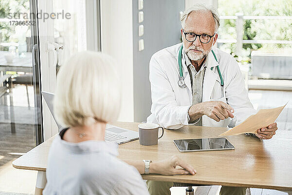 Männlicher Arzt hält einen medizinischen Bericht und betrachtet eine Patientin am Schreibtisch