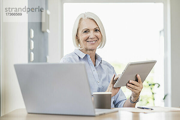 Lächelnde Geschäftsfrau  die ein digitales Tablet am Schreibtisch im Heimbüro hält