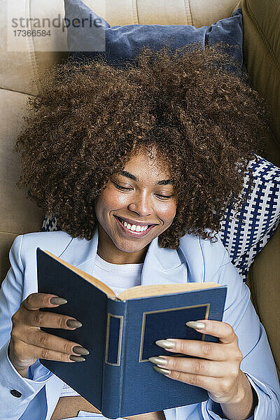 Lächelnde junge Geschäftsfrau im Blazer  die auf dem Sofa liegend ein Buch liest