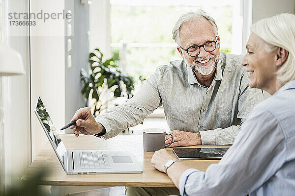 Lächelnder männlicher Berufstätiger  der auf einen Laptop zeigt  während er einer weiblichen Kollegin im Heimbüro etwas erklärt