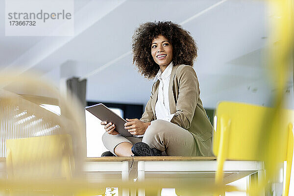 Afro-Geschäftsfrau hält digitales Tablet  während sie im Schneidersitz im Büro sitzt