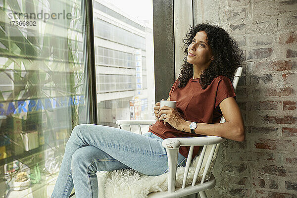 Nachdenkliche Frau mit Kaffeetasse auf einem Stuhl am Fenster sitzend
