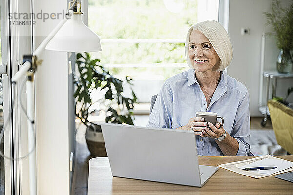 Lächelnde Geschäftsfrau  die wegschaut und eine Tasse im Büro zu Hause hält