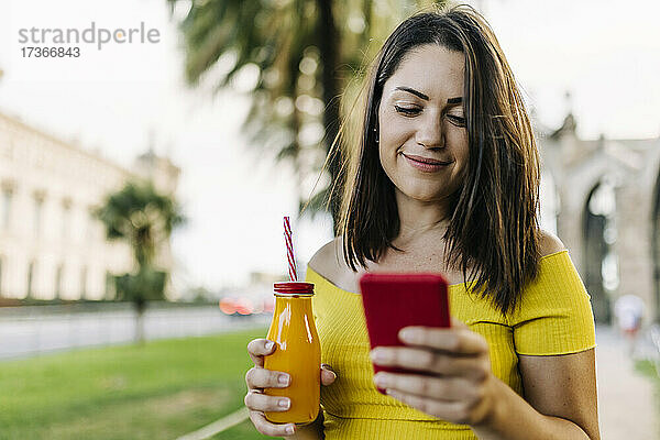 Lächelnde Frau  die eine Saftflasche hält und ein Smartphone benutzt