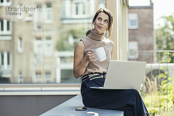 Nachdenkliche Geschäftsfrau sitzt mit Laptop und Kaffeetasse auf einer Stützmauer
