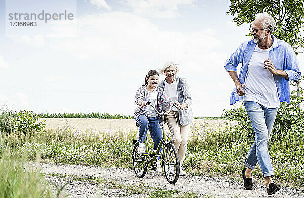 Mädchen fährt Fahrrad mit Frau hinter Mann auf unbefestigtem Weg