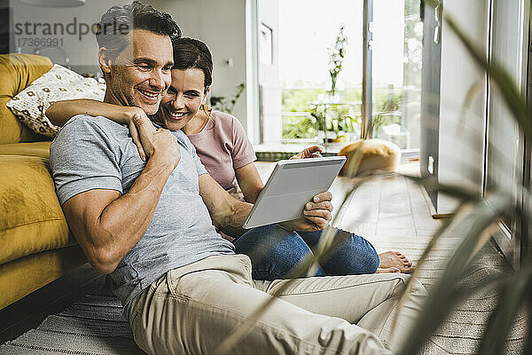 Lächelnde Frau mit einem Arm um einen Mann  der auf ein digitales Tablet schaut  während er zu Hause sitzt
