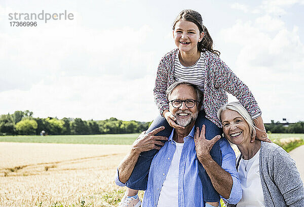 Fröhlicher Mann  der seine Enkelin auf der Schulter trägt  während die Frau mit ihm einen sonnigen Tag verbringt