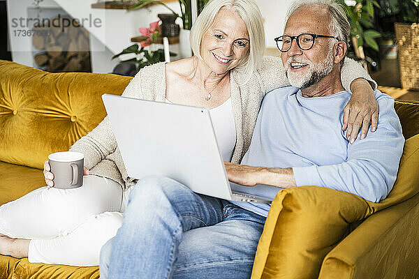 Lächelnde reife Frau  die den Arm um einen Mann legt  der zu Hause einen Laptop benutzt