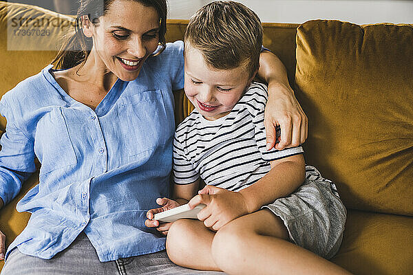 Frau mit Arm um Sohn  der auf dem Sofa zu Hause sein Smartphone benutzt