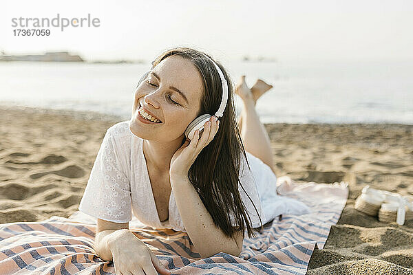 Lächelnde Frau hört Musik über Kopfhörer  während sie auf einem Handtuch liegt