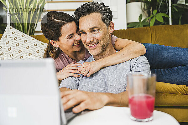 Lächelnde Frau  die einen Mann umarmt  der ein digitales Tablet zu Hause benutzt
