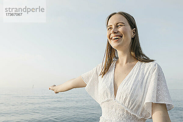 Schöne junge Frau  die lachend mit ausgestrecktem Arm am Strand steht