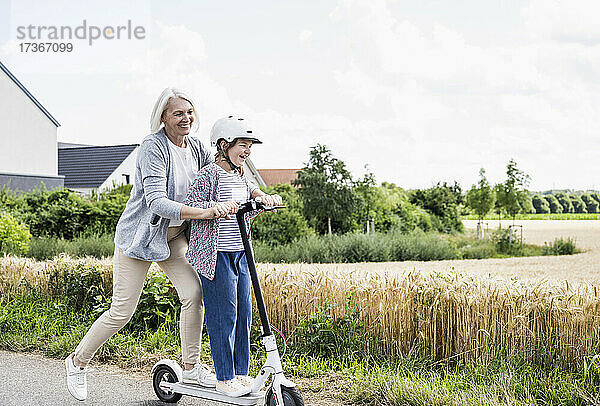 Lächelnde reife Frau  die mit ihrer Enkelin auf der Straße einen Motorroller fährt