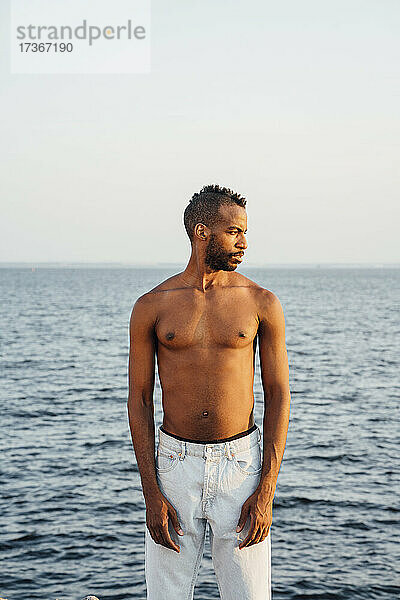 Mann ohne Hemd schaut weg  während er vor dem Meer steht