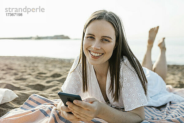 Lächelnde Frau mit Handy in der Hand beim Entspannen am Strand