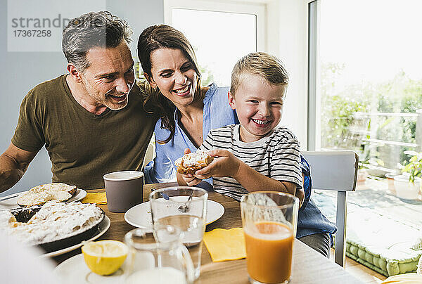 Lächelnder Junge hält Kuchen in der Hand  während er zu Hause mit der Familie frühstückt