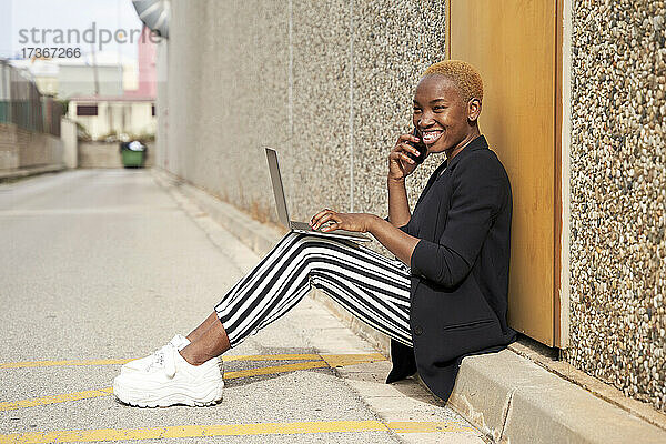 Lächelnde weibliche Fachkraft mit Laptop  die im Sonnenlicht am Telefon arbeitet