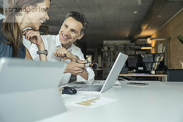 Männliche und weibliche Kollegen lächeln  während sie am Arbeitsplatz über einen Laptop diskutieren