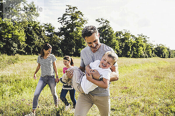 Lächelnder Vater trägt seinen Sohn beim Spaziergang mit der Familie auf einer Wiese