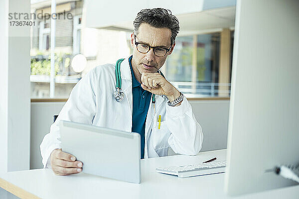 Männlicher Arzt  der ein digitales Tablet benutzt  während er im Büro sitzt