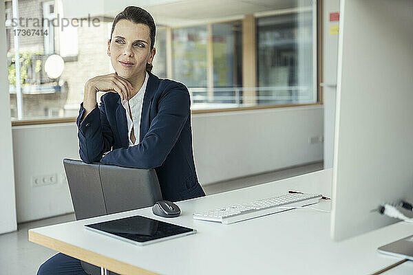 Nachdenkliche Geschäftsfrau mit der Hand am Kinn sitzt im Büro