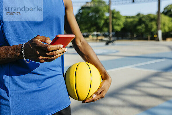 Mann  der ein Smartphone benutzt  während er einen Basketball auf einem Sportplatz hält