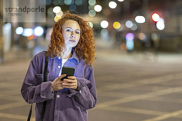 Rothaarige Frau mit Mobiltelefon  die nachts auf einer Kreuzung steht und wegschaut